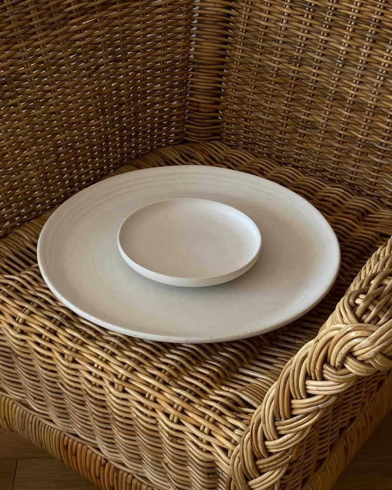 Großer weißer Servierteller aus Keramik mit kleinem weißen Keramikteller auf Korbstuhl.